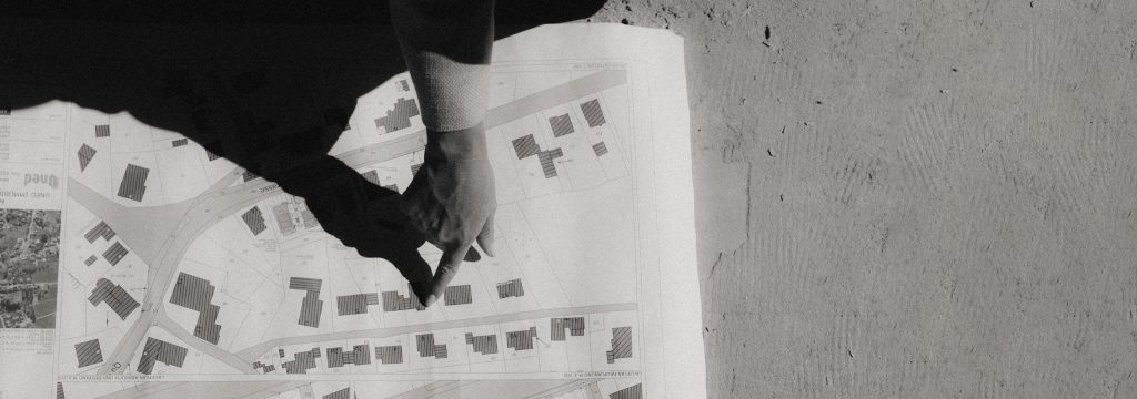 Eine Hand zeigt bei strahlendem Sonnenschein auf einen detaillierten Bauplan für ein Immobilienprojekt.